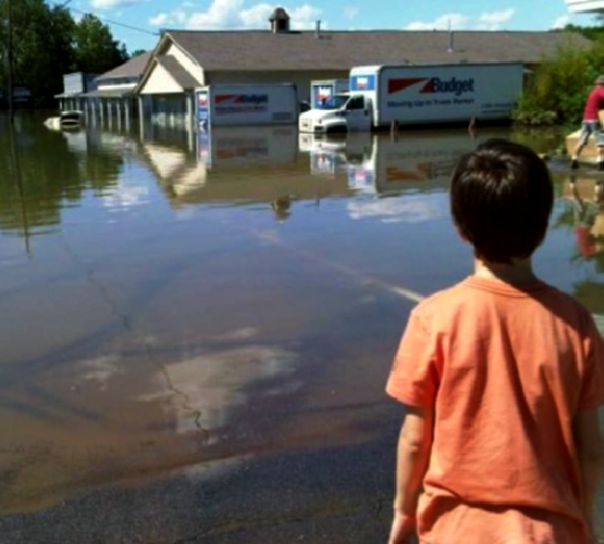 Parking lot of a shopping center submerged under flood water, with small boy surveying the damage.
